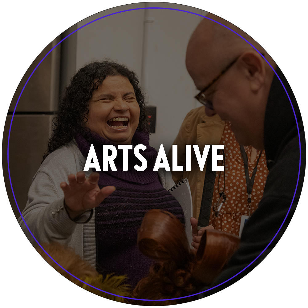 A circular image of a vision impaired woman with a large smile. She is being presented a costume piece to touch as part of Broadway Sacramento's Arts Alive tactile experience program, where guests can interact with props, costumes and production crew. Text in the center of the image reads "Arts Alive". 

You can click the image to redirect to the Arts Alive web page. 